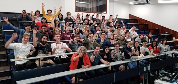 A bunch of sweaty nerds seated in a lecture theatre with their arms raised.